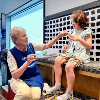 An elderly volunteer assists a young child in a colorful room, sharing a warm interaction. The environment is casual and friendly.