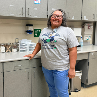 A smiling school nurse stands in a clinic with medical supplies organized on the countertop. The setting is calm and welcoming.