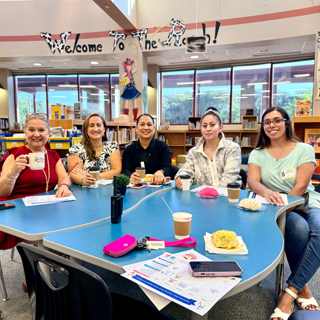 A group of five people sitting around a table in a library, enjoying coffee and pastries. The atmosphere is friendly and welcoming.
