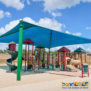 A vibrant playground with a large sunshade featuring children playing on the structures under a bright blue sky. The atmosphere is lively and cheerful.