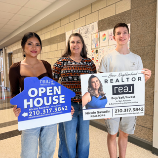 Three people smile while holding real estate promotional signs in a hallway. The signs advertise an open house by Realtor Nicole Savadin, showcasing a friendly and welcoming community spirit.