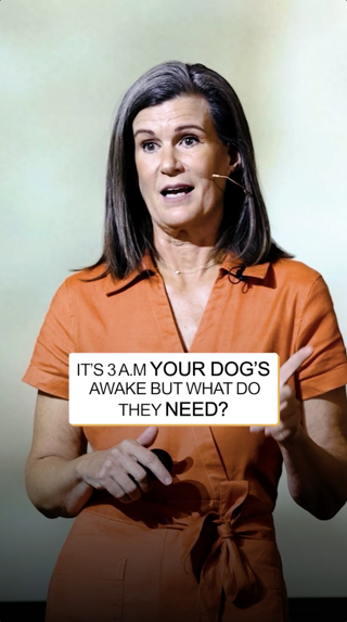 A woman in an orange jumpsuit speaks on stage with a microphone headset. The text overlay addresses dog behavior in the early hours.