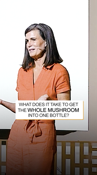 A woman in an orange outfit speaks on stage with a presentation asking about getting mushrooms into a bottle.
