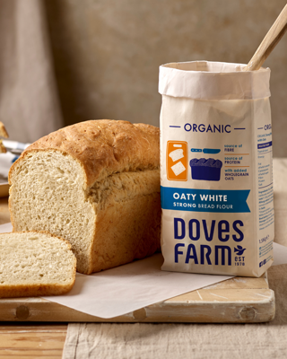 A freshly baked loaf of bread sits next to a bag of organic strong bread flour on a wooden board. The scene conveys a homemade, artisanal baking vibe.