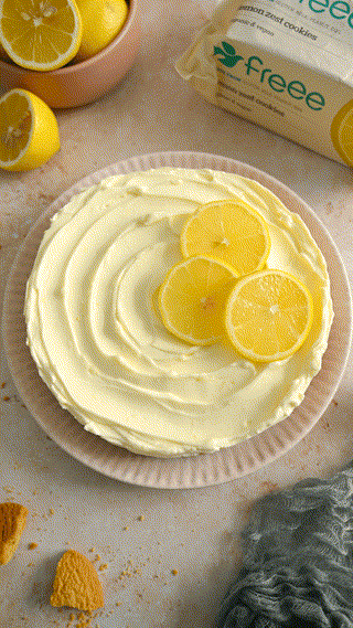A creamy lemon cake topped with fresh lemon slices. The cake is displayed on a plate with surrounding lemons and a packet labeled 'lemon zest cookies' in the background. The scene exudes a fresh and inviting atmosphere.