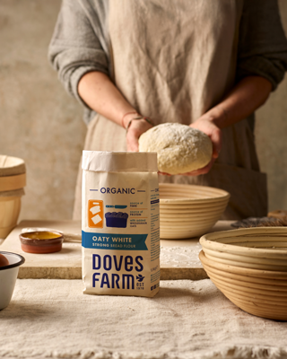 A person in an apron prepares dough with Doves Farm organic flour displayed in a rustic setting. The mood is warm and artisanal.