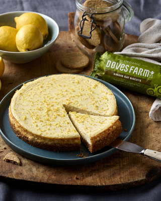 A delicious lemon cheesecake garnished with lemon zest, served on a wooden board. Accompanied by lemons, biscuits, and a package of organic ginger oat biscuits.