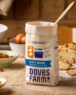 A bag of Doves Farm Oaty White Strong Bread Flour on a wooden table. The scene has a warm, rustic kitchen vibe.