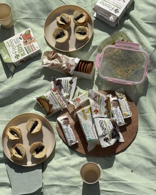 A bright outdoor picnic setup featuring a variety of gluten-free snacks, muffins, and tea on a pastel green blanket. The scene conveys a relaxed and inviting mood.