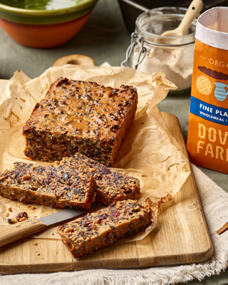 A freshly baked fruitcake with visible dried fruits, sliced on a wooden board next to a bag of organic flour. The scene is cozy and inviting, perfect for a rustic kitchen setting.
