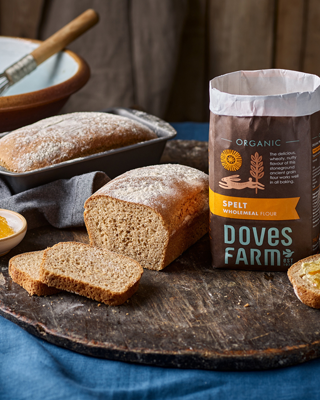 A loaf of homemade organic spelt bread is placed on a rustic board, alongside a bag of Doves Farm Spelt Wholemeal Flour, showcasing a warm and inviting atmosphere.