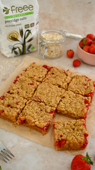 Gluten-free strawberry oat bars on parchment paper with fresh strawberries and a bag of porridge oats. The image conveys a homemade, healthy dessert vibe.