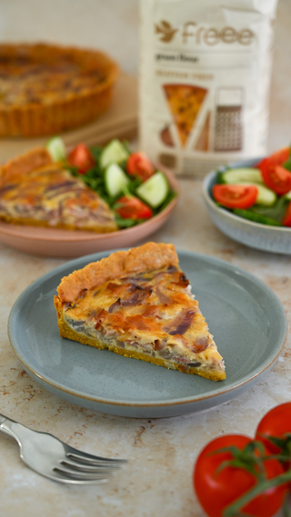 A slice of savory quiche is presented on a light blue plate, accompanied by a fresh salad of cucumbers and tomatoes. The background shows a full quiche and gluten-free flour package, creating a homely and inviting culinary atmosphere.
