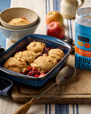 A homemade apple and berry cobbler baked in a blue dish, set on a rustic wooden board. Nearby are fresh apples and organic self-raising flour.