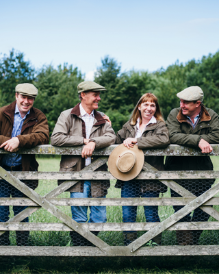 Four people in outdoor attire lean on a wooden farm gate, sharing a cheerful moment in a lush, green setting.