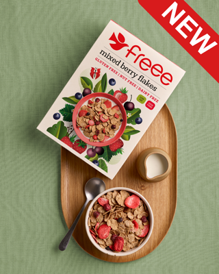 A top-down view of a 'new' gluten-free cereal box and a bowl of mixed berry flakes on a wooden tray with a green cloth background. Includes a small jug of milk and a spoon.