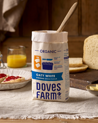 A package of organic oaty white bread flour from Doves Farm, placed on a table with bread slices and a glass of orange juice nearby. The scene conveys a cozy, homemade baking vibe.