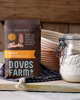 A bag of Doves Farm organic spelt wholemeal flour displayed with baking supplies in a rustic kitchen setting. The image conveys a warm and natural ambiance, ideal for bakers.