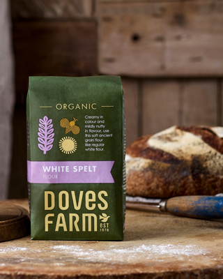 A package of organic white spelt flour from Doves Farm sits on a rustic wooden surface, with a fresh loaf of bread in the background. The packaging features a green and purple color scheme, emphasizing its organic quality.