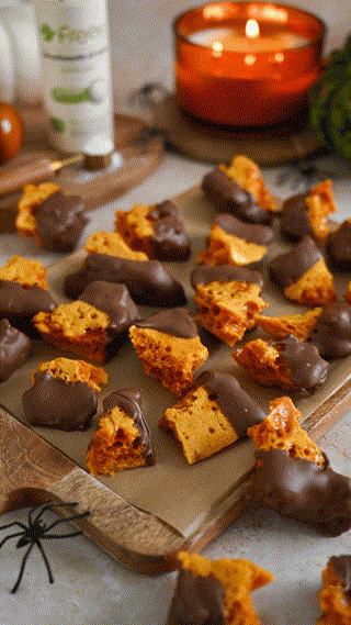 A tray of chocolate-dipped honeycomb candy pieces displayed on parchment paper. A warm, cozy ambiance is created by an orange candle and small decorative spiders, suggesting a Halloween theme.