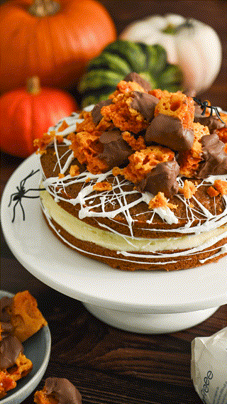 A Halloween-themed cake decorated with honeycomb pieces and chocolate, adorned with spider web icing. Pumpkins in the background add a festive touch.