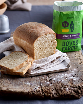 A rustic scene featuring a loaf of freshly baked bread with slices arranged in front and a bag of organic strong white bread flour in the background. The setting is cozy and homely, with a wooden surface and soft cloth.