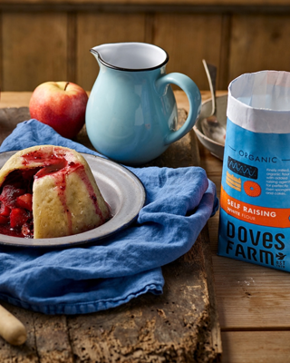 A rustic setting featuring a steamed pudding with fruit, a blue pitcher, an apple, and a bag of Doves Farm flour.