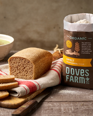 A rustic scene showcasing wholemeal spelt bread on a wooden board, accompanied by a bag of organic spelt flour. The warm, inviting setup emphasizes the natural, wholesome quality of the products.