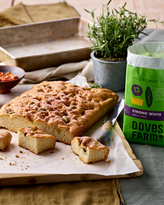 A delicious homemade focaccia bread sits on a parchment-lined board, accompanied by a bag of flour and rosemary. The scene conveys a rustic kitchen vibe.
