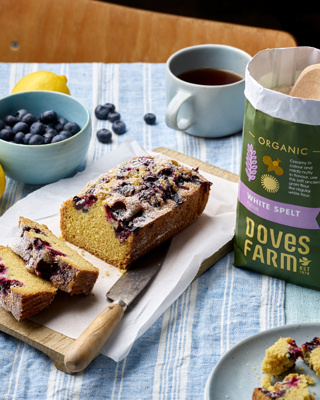 A freshly baked blueberry loaf sits on a wooden board, surrounded by a cup of tea, blueberries, and a bag of organic white spelt flour. The setting conveys a cozy and inviting atmosphere.