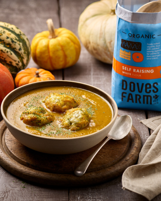 A cozy autumn scene with a bowl of pumpkin soup topped with herb dumplings. Surrounded by pumpkins and a bag of flour on a rustic wooden table.
